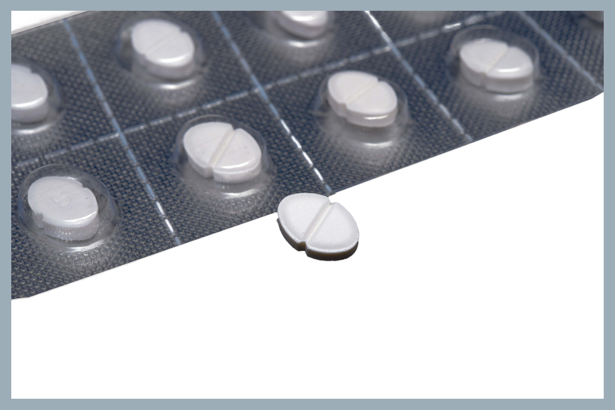 White tablet in front of blister pack of tablets; it is oval-shaped and has an indented line in the centre